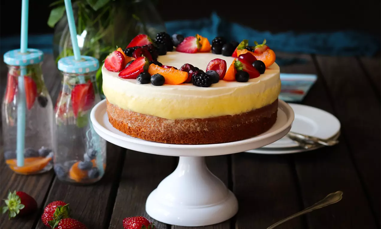 Svetložltá ovocná torta na svetlom podstavci s vrstvami krémovej plnky, ozdobená čerstvými jahodami, malinami, černicami a marhuľami