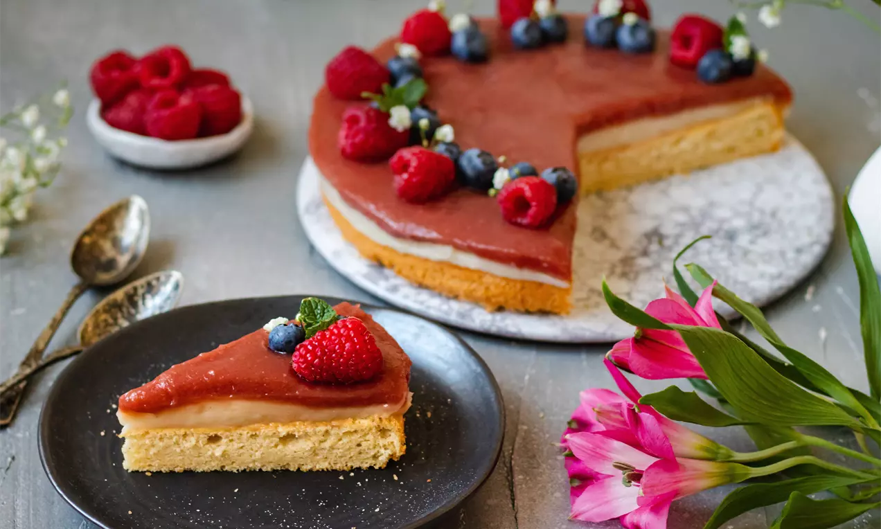 Torta s jemným dvojfarebným krémom na zlatistom korpuse, ozdobená malinami a čučoriedkami, podávaná na tanieriku s kvetmi a lyžičkami