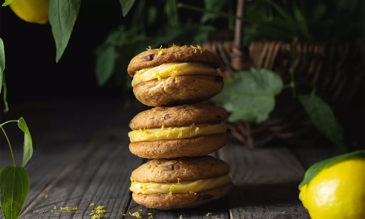 Tri okrúhle hnedasté cookies s viditeľnými kúskami čokolády, zlepené žltým krémom, poukladané na sebe na tmavom drevenom stole s citrónmi a zeleňou v pozadí