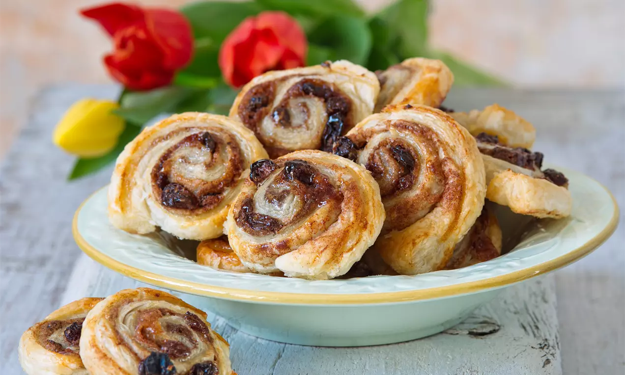 Vanilkovo-škoricové slimáky so zlatistou špirálovou štruktúrou, jemne lesklé, položené na pastelovom tanieri s červenými a žltými tulipánmi
