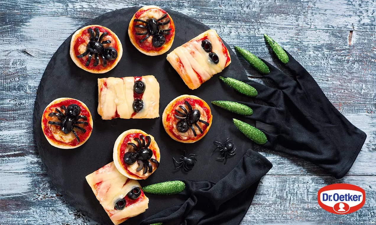 Mini pizze na tenkom čiernom tanieri, okrúhle a hranaté, s červenou paradajkovou omáčkou, roztopeným syrom a olivovými pavúkmi, obklopené čiernou rukavicou a zelenými sladkosťami