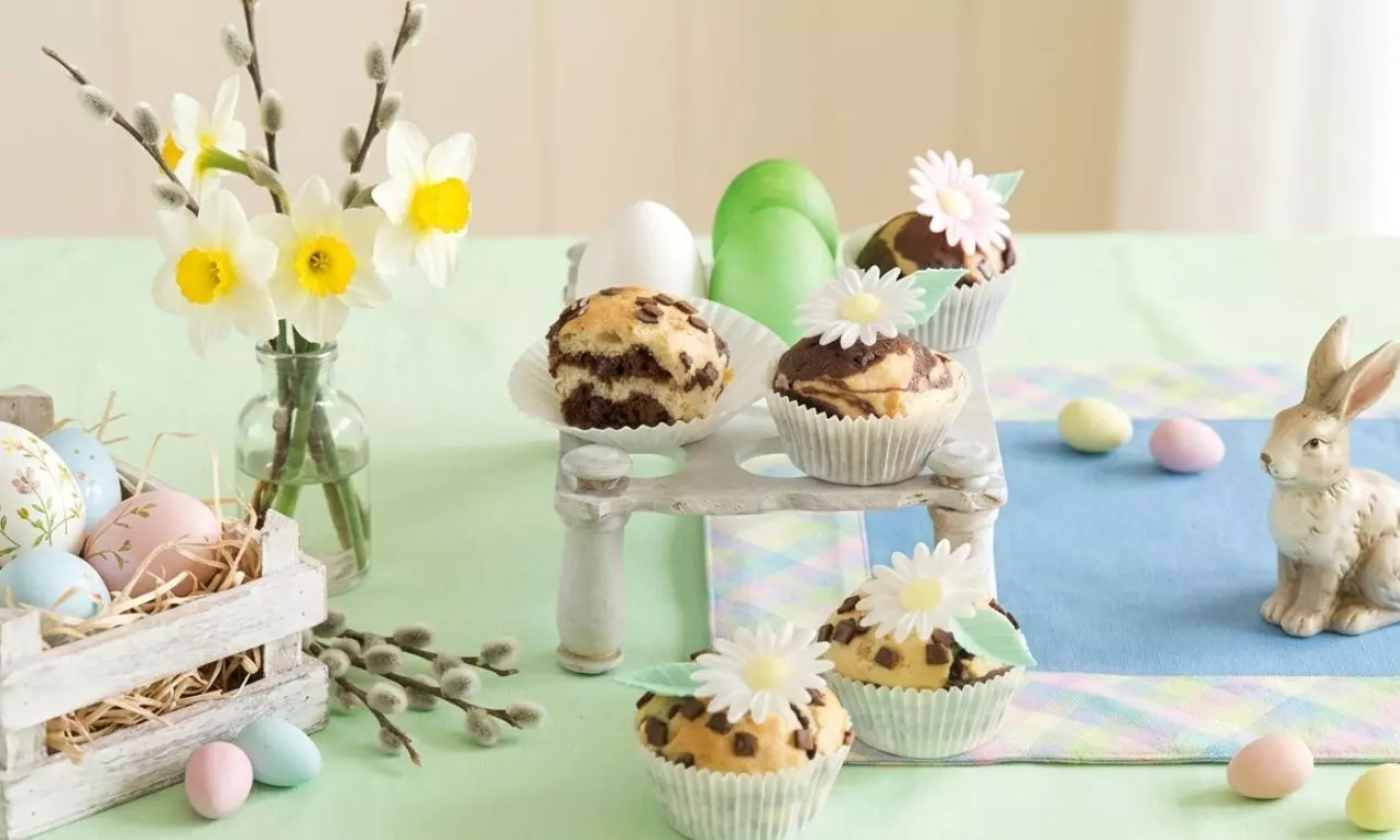 Strakaté muffiny vo svetlých košíčkoch, kakaovo-vanilkové, zdobené bielymi margarétkami a zelenými lístkami na pastelovom prestieraní