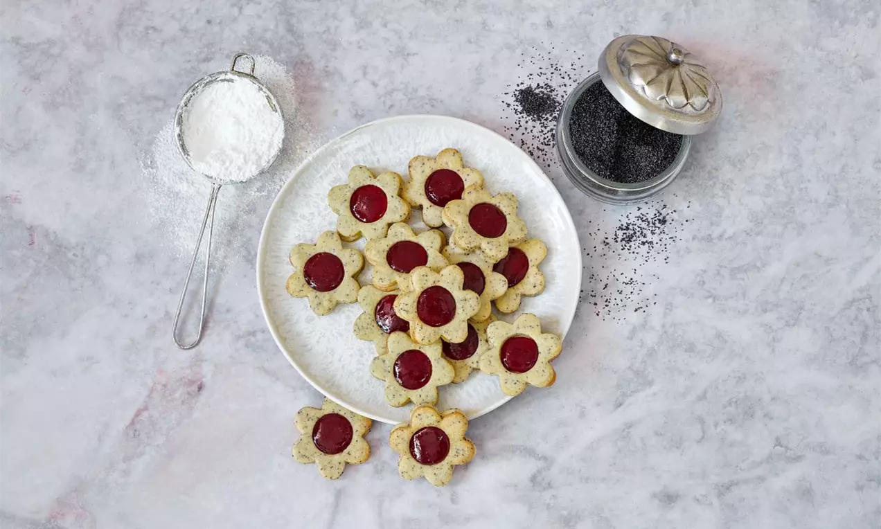 Makové linecké kvietky svetlo zlatej farby s červeným stredom usporiadané na bielom tanieri, vedľa sitka s práškovým cukrom a pohár maku