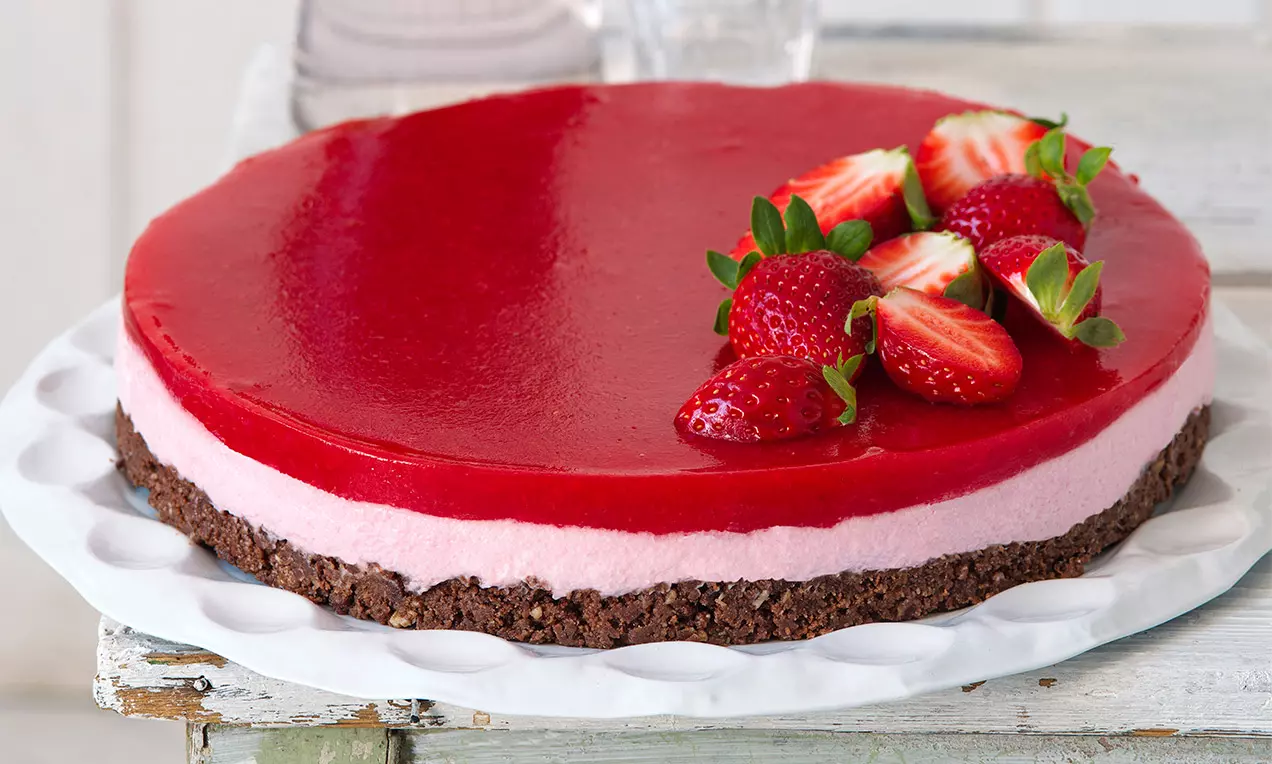 Jahodový cheesecake s červenou lesklou polevou, vrstvou ružového krému, hnedým sušienkovým základom, ozdobený čerstvými jahodami na bielom tanieri