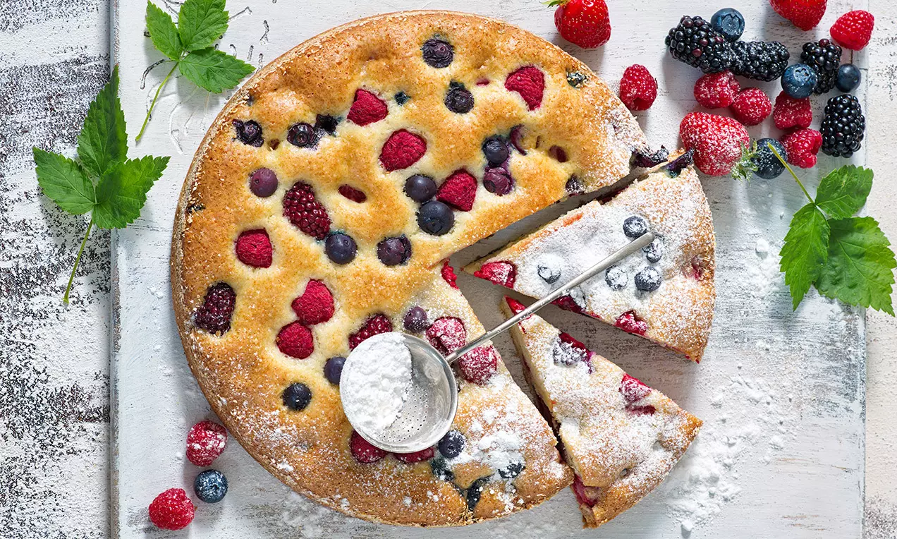 Tortová bublanina svetlo zlatistá s napečenými malinami, černicami a čučoriedkami, posypaná práškovým cukrom, na drevenom podnose
