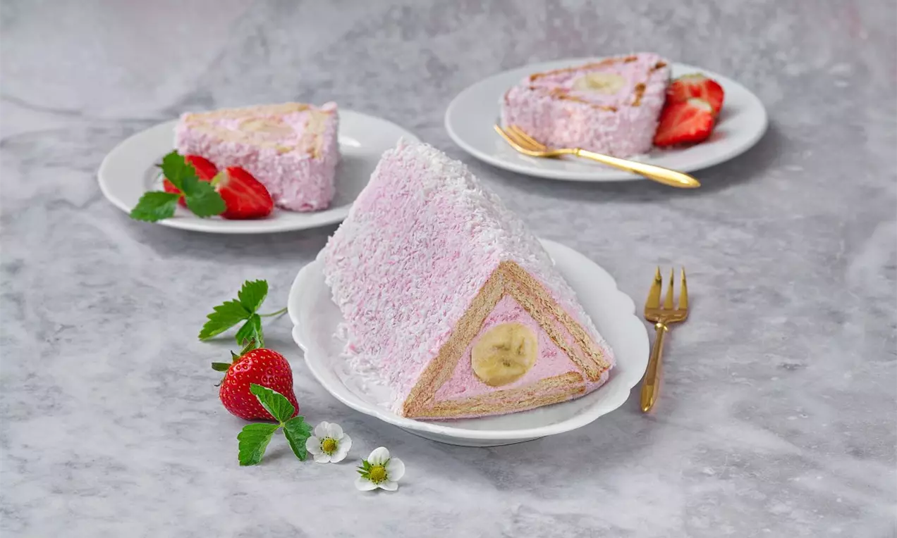 Trojuholníkový rez s ružovou jahodovou náplňou, ukrytou v kokosovej posypke, uprostred banán, podávaný na bielom tanieri s čerstvými jahodami