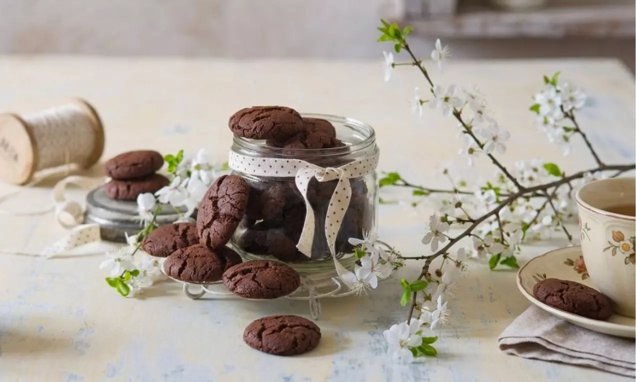 Tmavohnedé kakaové cookies s popraskaným povrchom, rozložené vedľa sklenenej nádoby ozdobenej stuhou, obklopené kvitnúcimi vetvičkami
