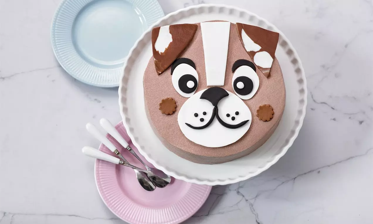 Tvarohová torta okrúhleho tvaru s tvárou psa z hnedej, bielej a čiernej polevy na bielom tanieri, vedľa farebných tanierikov a lyžičiek