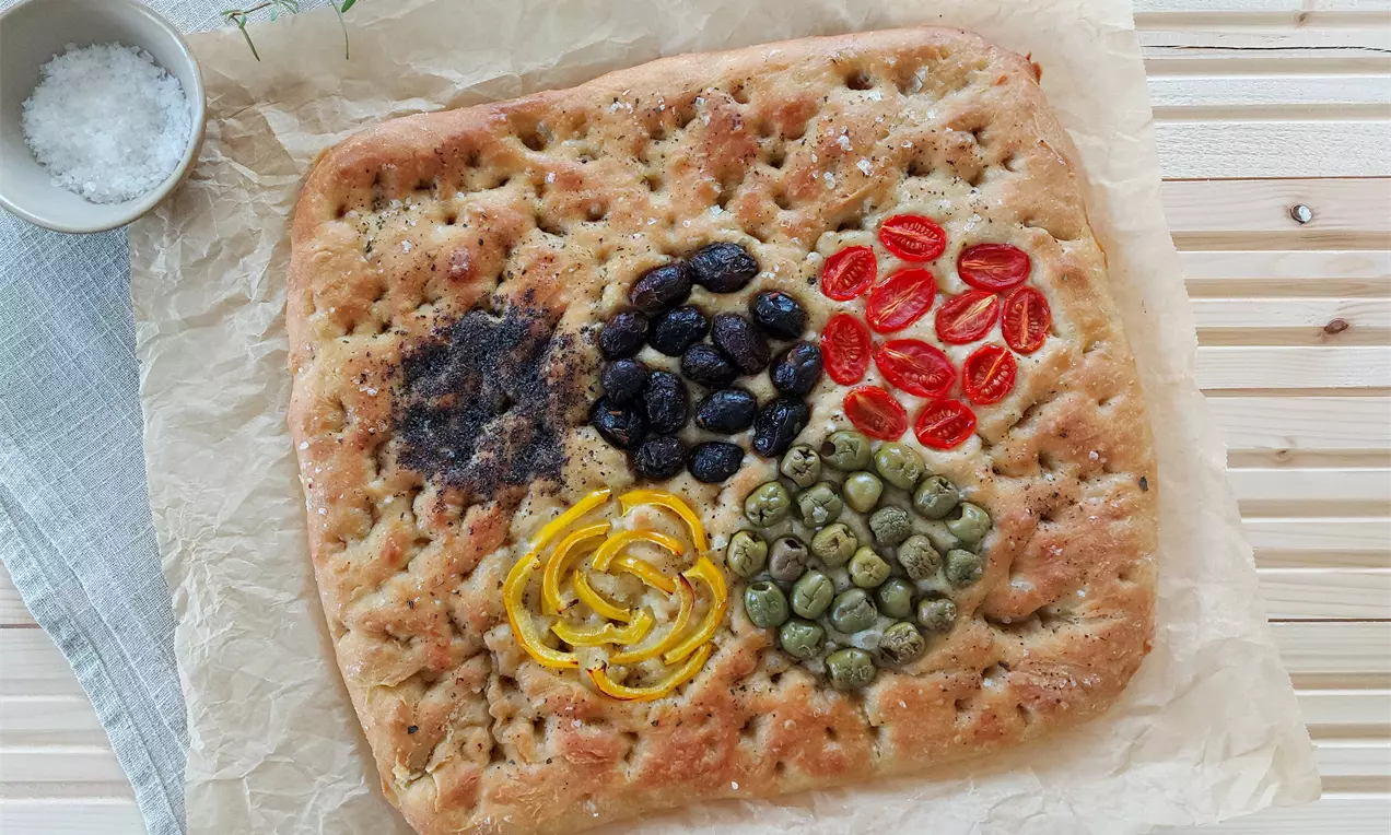 Zlatistá focaccia v obdĺžnikovom tvare, ozdobená farebným usporiadaním čiernych olív, červenej rajčiny, zelených kapár a žltých citrusových kúskov na papieri na pečenie