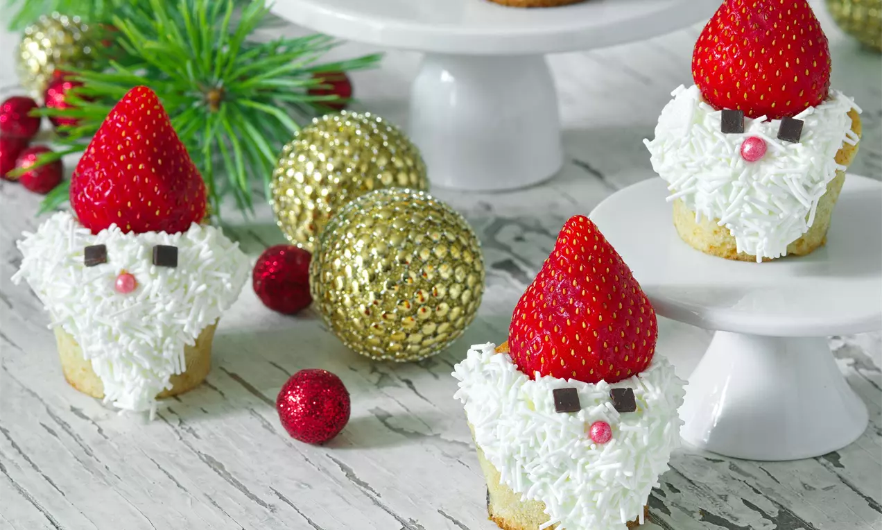 Muffiny s bielou snehovou polevou, červené jahodové čiapky, čokoládové okuliare a cukríkové nosy usporiadané na podnosoch s vianočnými ozdobami