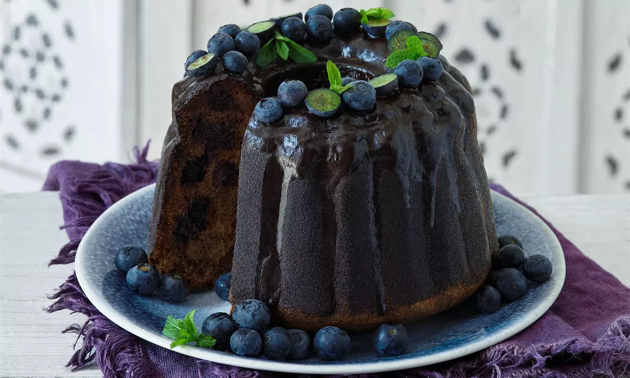 Kakaová bábovka plnená čučoriedkami, zdobená čerstvými čučoriedkami a lístkami čučoriedok na bielom podklade a fialovej šatke.