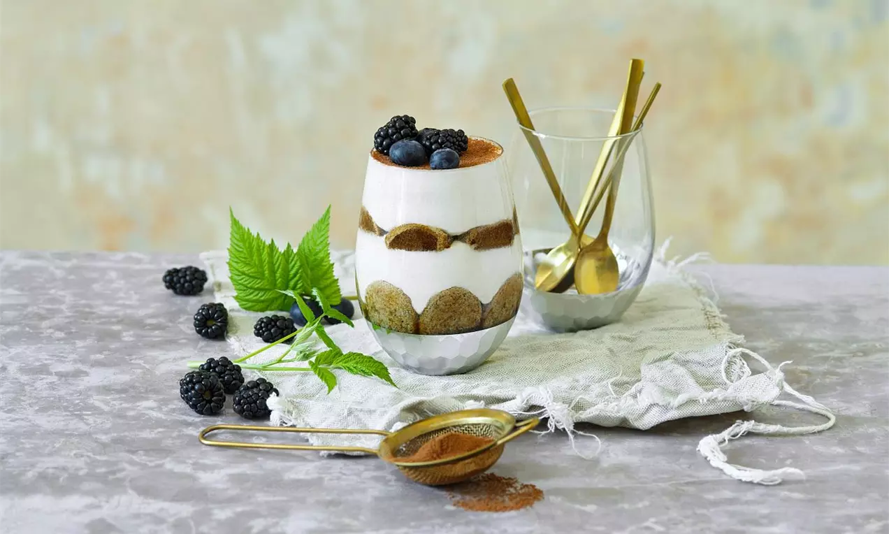 Poháre s vrstveným dezertom z krémového bieleho mascarpone, kakaového prášku, piškót, černíc a čučoriedok na šedom povrchu