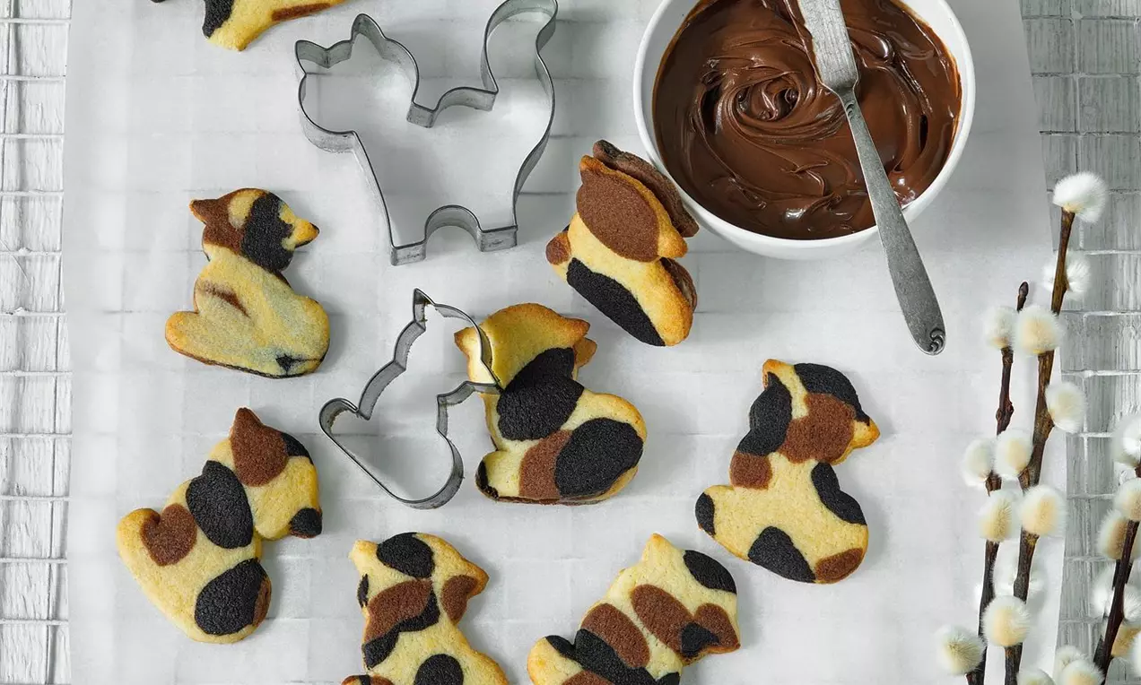 Sušienky v tvare mačičiek s mramorovým vzorom v odtieňoch žltej, hnedej a čiernej, položené na bielom podklade vedľa formičiek a misky čokolády