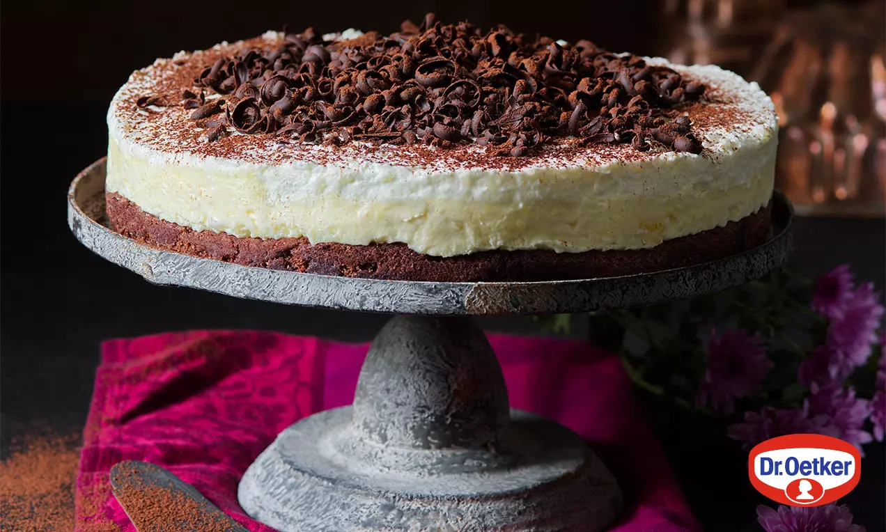 Torta s krémovou svetlou plnkou na tmavom kakaovom základe, posypaná bohatými hoblinkami čokolády na rustikálnom podstavci