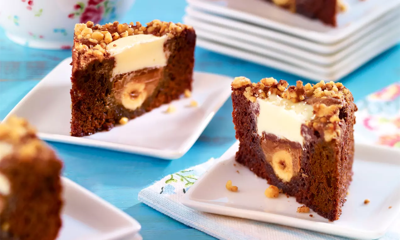 Trojuholníky tmavohnedého brownies s jemnou textúrou, svetlou krémovou vrstvou a posypané nadrobno nasekanými orechmi na bielych tanieroch