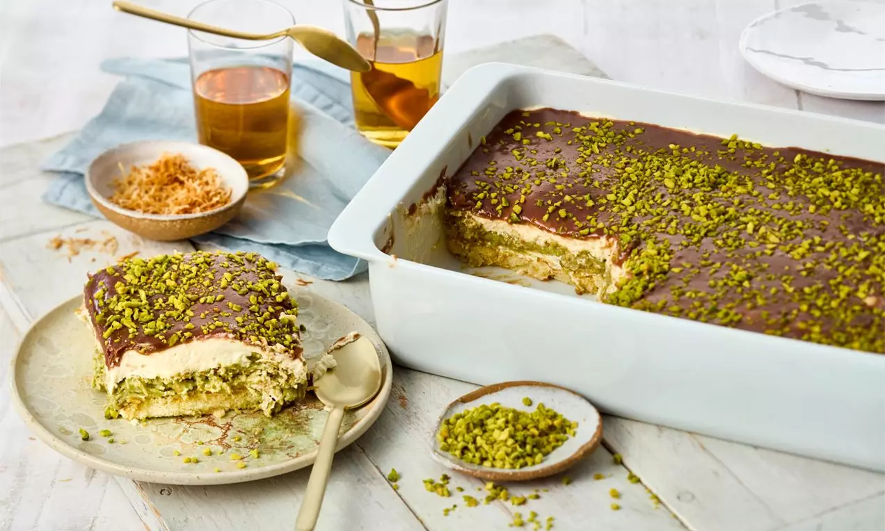 Dubajské čokoládové tiramisu s vrstvou lesklej tmavej čokolády, posypané zelenými pistáciami, podávané na tanieri vedľa zapekacej misy