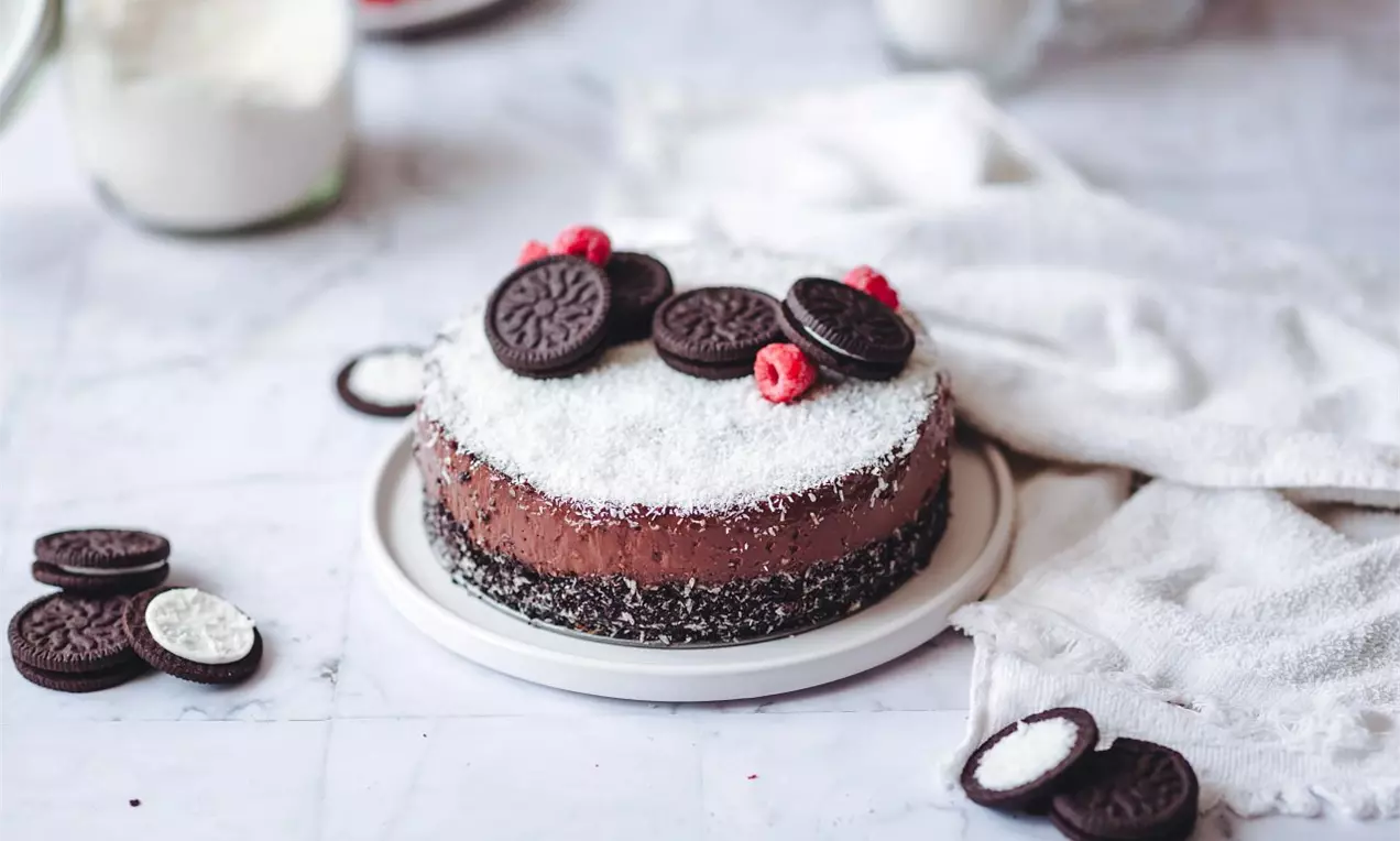 Okrúhly čokoládovo-kokosový cheesecake sypaný strúhaným kokosom, zdobený sušienkami Oreo a malinami na bielom tanieri na mramorovom podklade