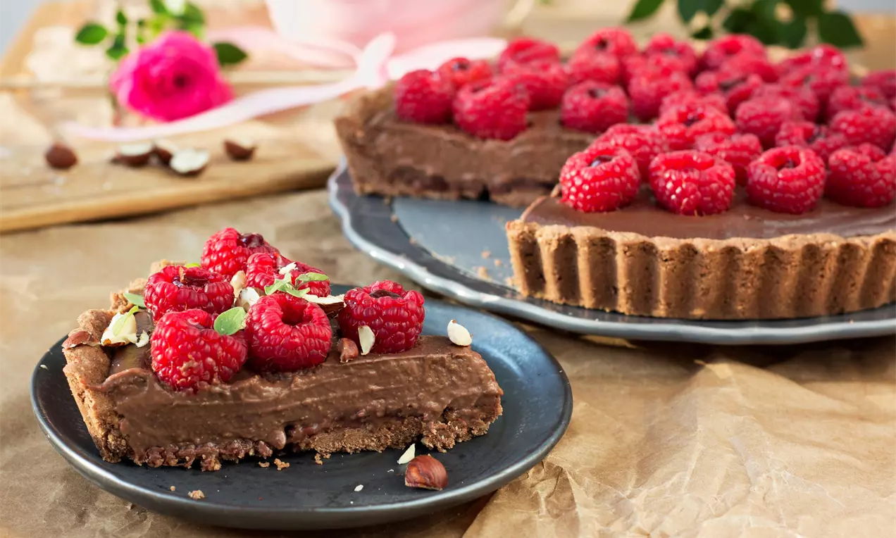 Čokoládový BB tart s hladkým lesklým kakaovým povrchom zdobený šťavnatými tmavočervenými malinami a kúskami orieškov na tmavom tanieri