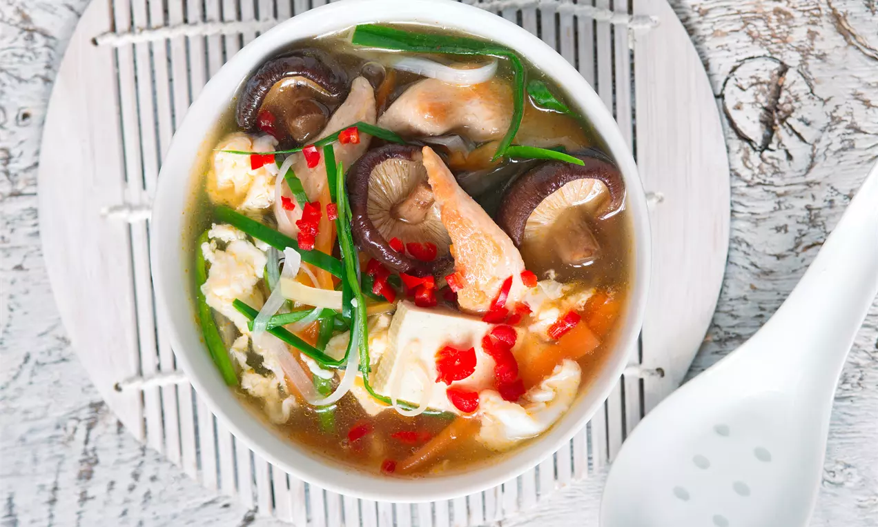 Svetlohnedý vývar s kúskami bieleho tofu, tmavohnedých húb, červených chilli a zelených cibuľových plátkov v bielej miske na bambusovej podložke