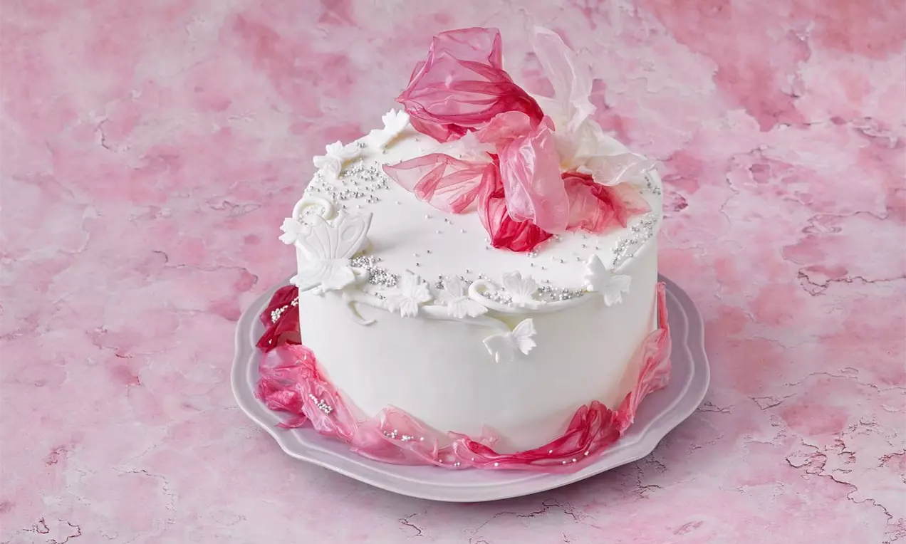 Biela torta s hladkým povrchom, ozdobená ružovými a bielymi kvetinovými dekoráciami na svetlom tanieri, položená na ružovom mramorovom pozadí