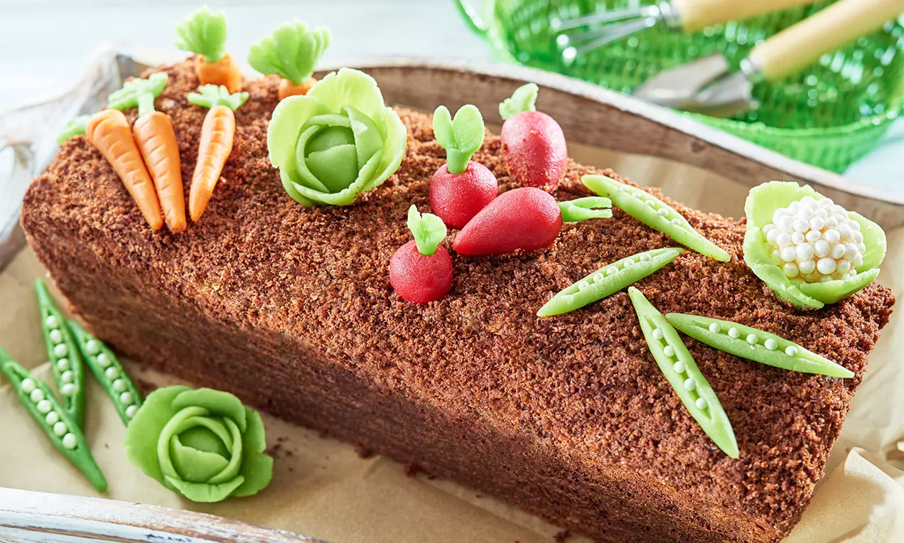 Podlhovastá torta s hnedým drobivým povrchom, ozdobená farebnými marcipánovými zeleninami v podobe mrkvy, kapusty, hrášku a reďkoviek