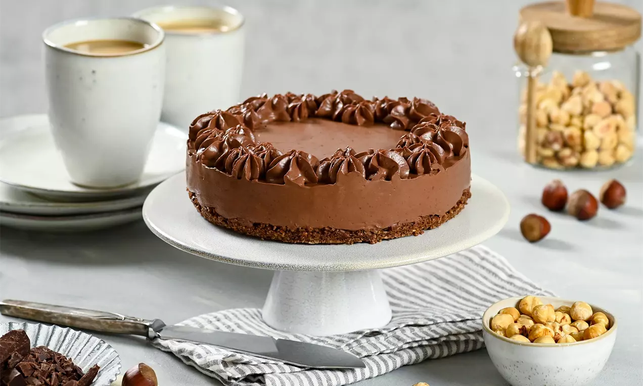 Okružná vegánska čokoládová torta v tmavohnedom odtieni s hladkou textúrou, zdobená výraznými krémovými špirálami na bielom stojane