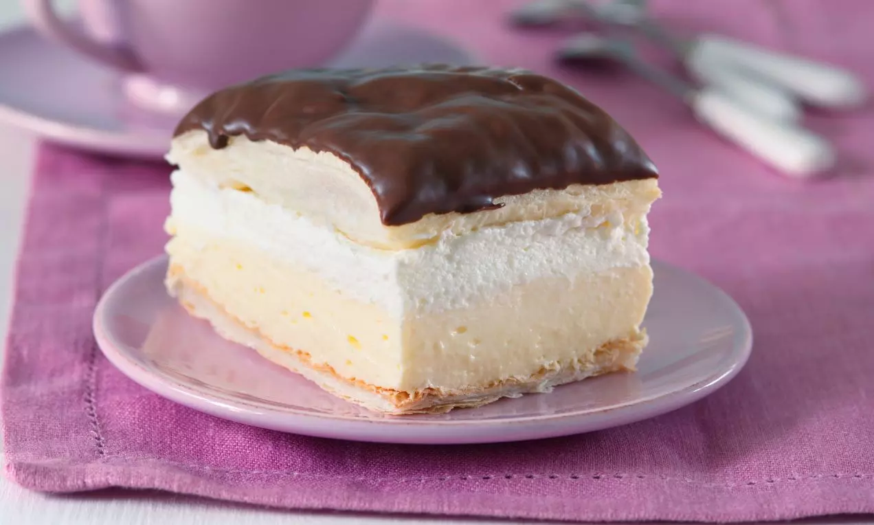 Dvojvrstvový zákusok s krémovým a šľahačkovým jadrom, poliaty lesklou čokoládovou polevou na ružovom tanieriku s pastelovým pozadím