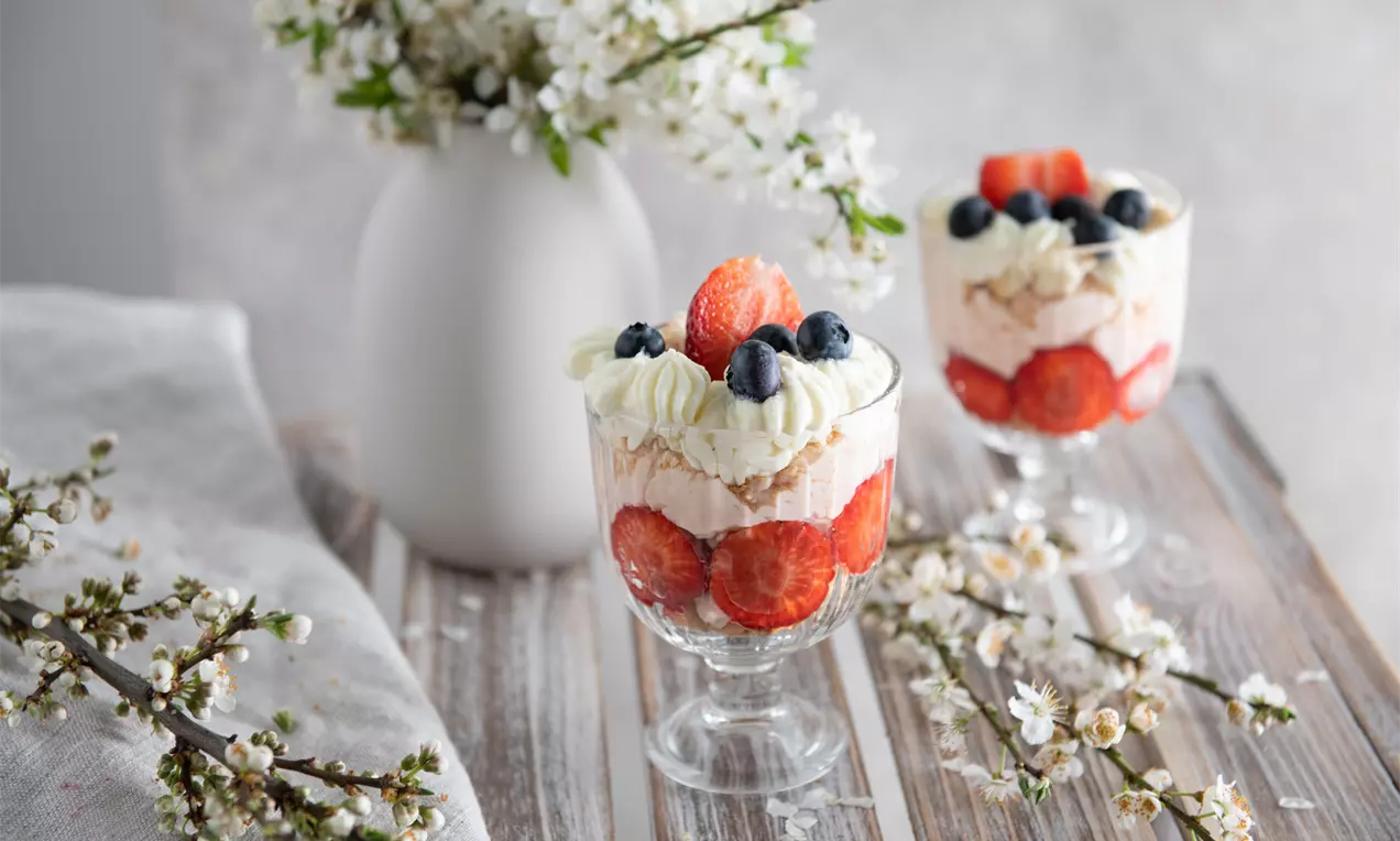 Jemná vrstva mascarpone krému a piškót, obklopená plátkami čerstvých červených jahôd, ozdobená čučoriedkami a šľahačkou v sklenenom pohári