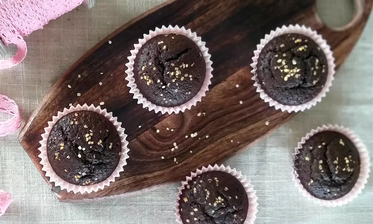 Tmavohnedé okrúhle muffiny v bledoružových košíčkoch na drevenej doske, posypané drobnými semienkami, usporiadané nepravidelne