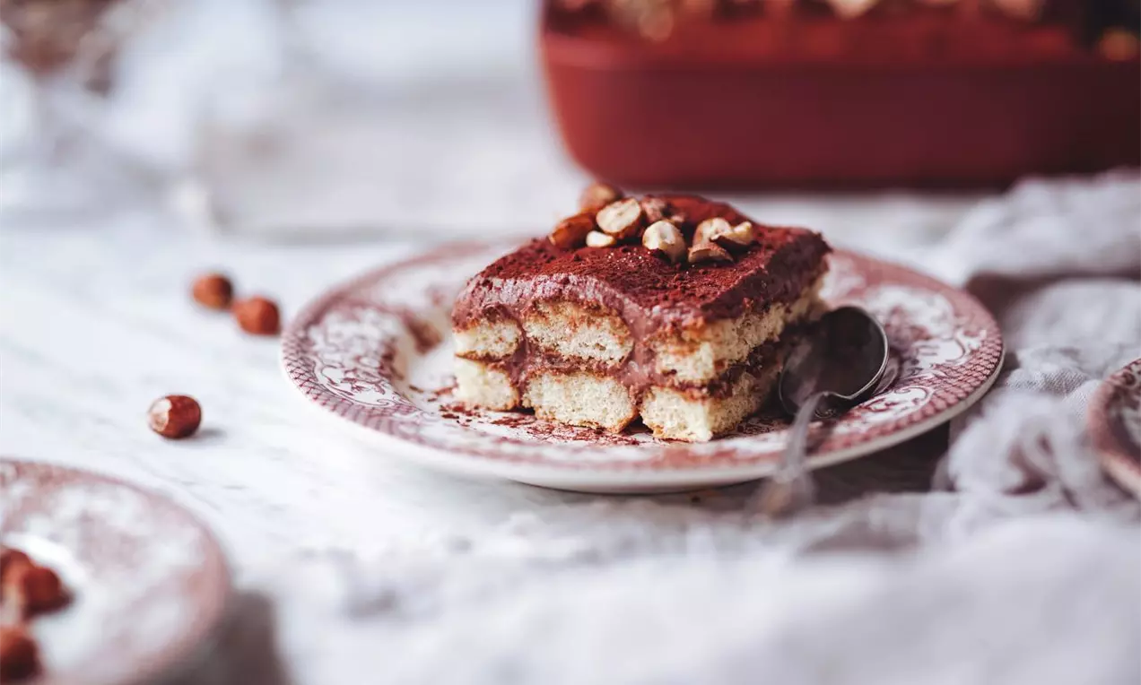 Vrstvený dezert prekladaný piškótami, krémovou vrstvou sypaný kakaom a ozdobený orechmi na zdobnom dezertnom tanieriku a mramorovej doske