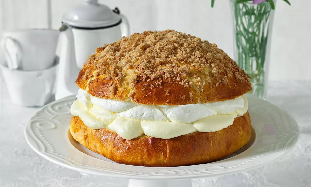 Pražský koláč zlatohnedej farby s nadýchaným kysnutým cestom, vrstvou bielej šľahačky a drobenkou na elegantnom bielom podnosovom tanieri