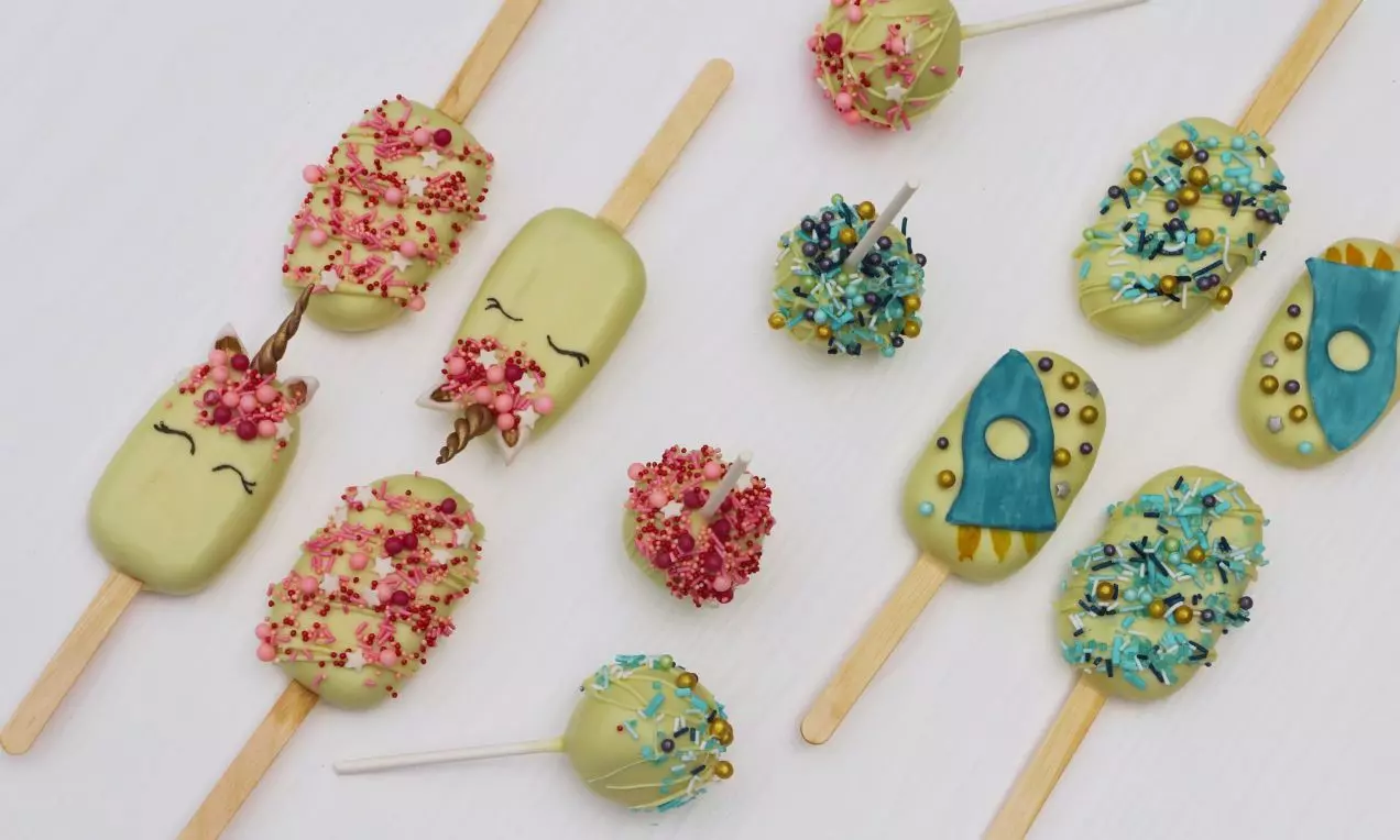 Banánovo-kakaové nanuky svetlej zelenej farby na drevených paličkách, zdobené červenými, modrými a zlatými prísadami, vedľa cakepops na bielom pozadí