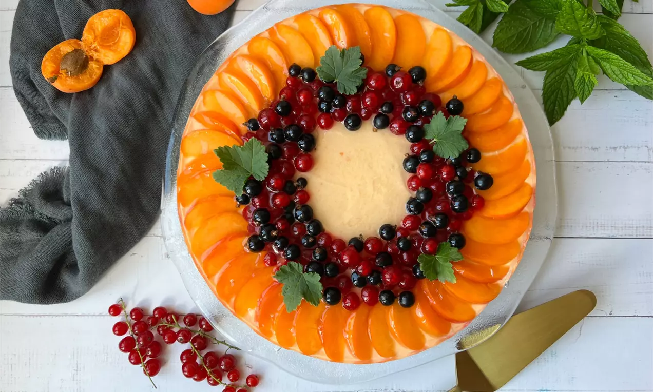 Ovocná torta s hladkým svetlým krémom, ozdobená krúživými radmi oranžových marhúľ a farebných bobúľ so zelenými lístkami mäty