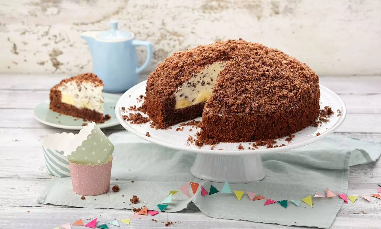 Okrúhla torta z tmavého kakaového cesta s kopulou z drvených omrviniek, plnená svetlým krémom a banánmi, na bielom podnose