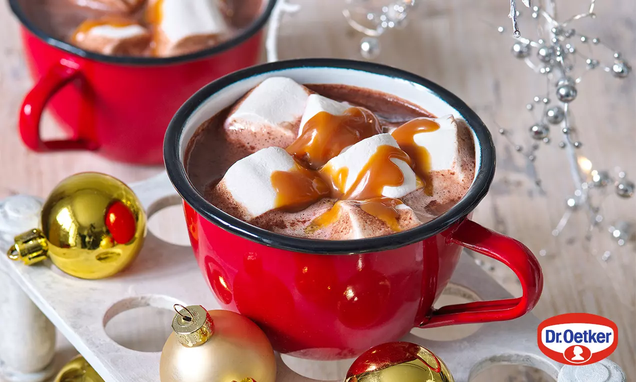 Karamelová horúca čokoláda v červenom hrnčeku, ozdobená nadýchanými bielymi marshmallows a prúžkami zlatistého karamelu