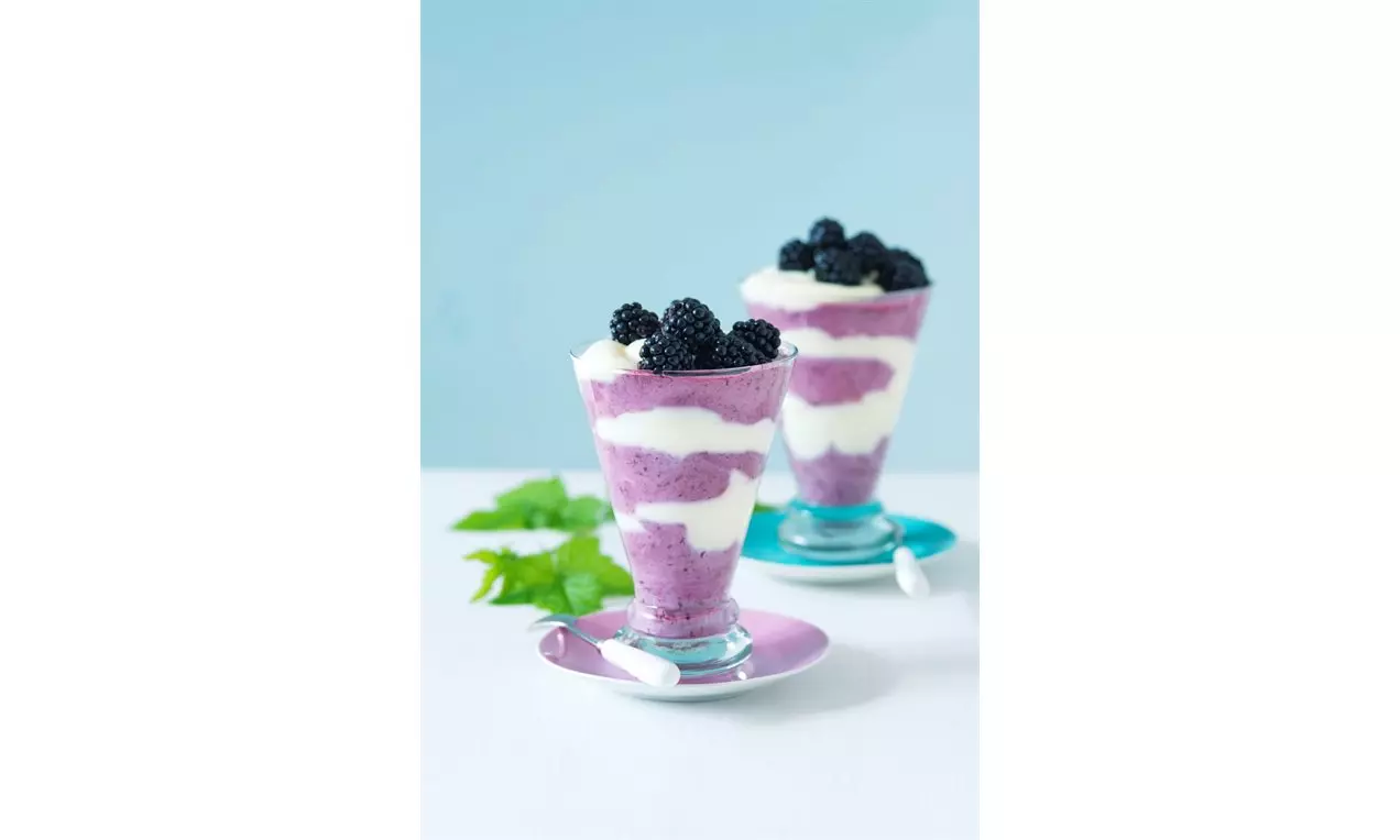 Vrstva krémového vanilkového pudingu bielej farby a fialového ovocného pyré ozdobená čerstvými černicami v sklenenom pohári na podšálke