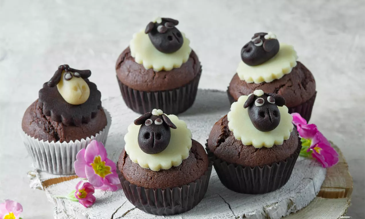 Kakaové muffiny tmavohnedé, s detailne modelovanými ovečkami z čokolády a bieleho fondánu, na drevenom podnose, obklopené ružovými kvetmi