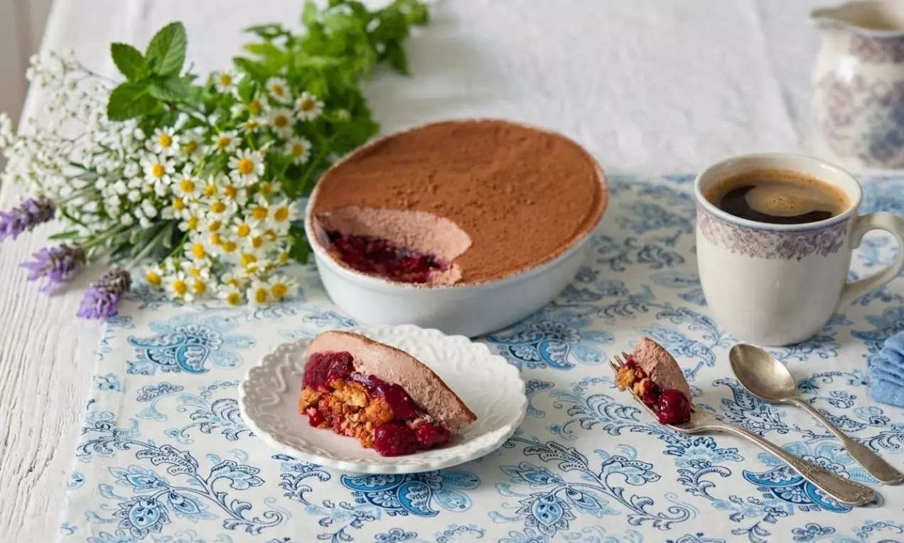 Tvarohové tiramisu s jemným kakaovým povrchom, vrstvou čerešňovej plnky a krehkým základom na bielom tanieri vedľa šálky kávy
