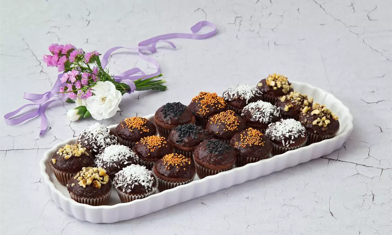 Tmavohnedé mini muffiny s rôznymi posypmi, ako sú kokos, mak, karamel a orechy, na porcelánovom tanieri vedľa dekoratívnej kvetinovej väzby