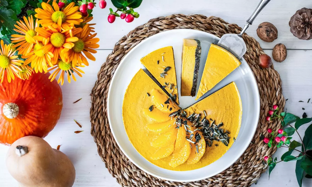 Nakrojený žltý cheesecake na bielom tanieriku a prútenom prestieraní, zdobený tekvicovými semienkami a filetami z pomaranča, s okolitou dekoráciou čerstvých tekvíc a jesenných kvetov.