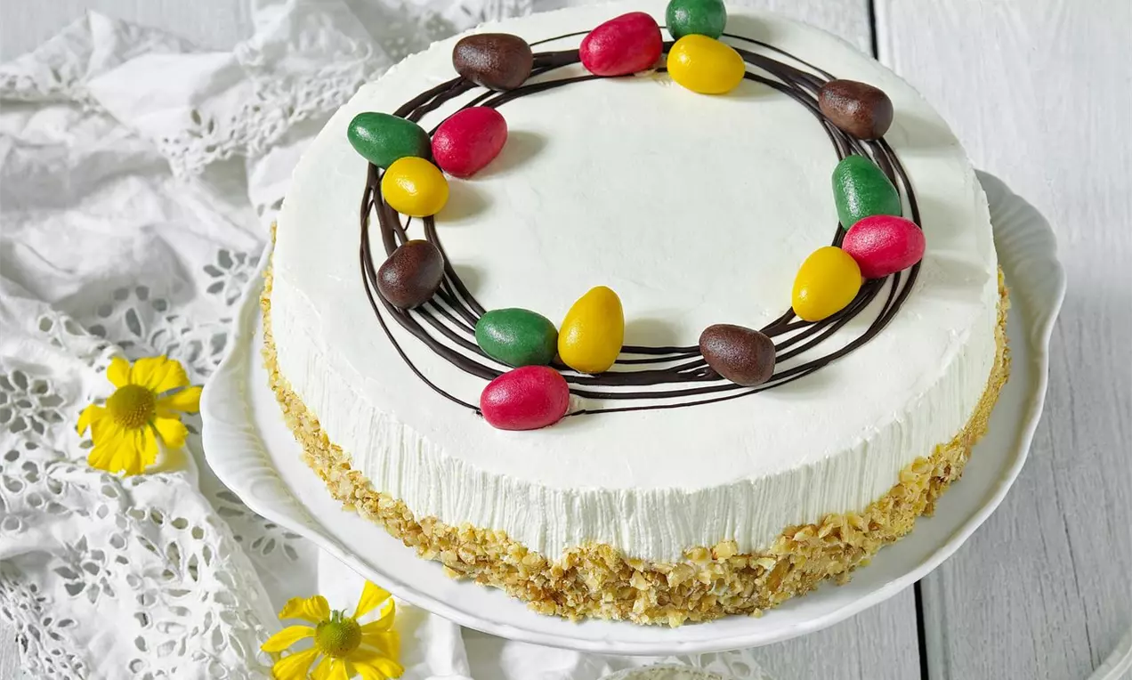 Krémová torta s jemným banánovým nádychom na chrumkavom orechovom základe, zdobená farebnými vajíčkami a čokoládovým vencom na bielom tanieri