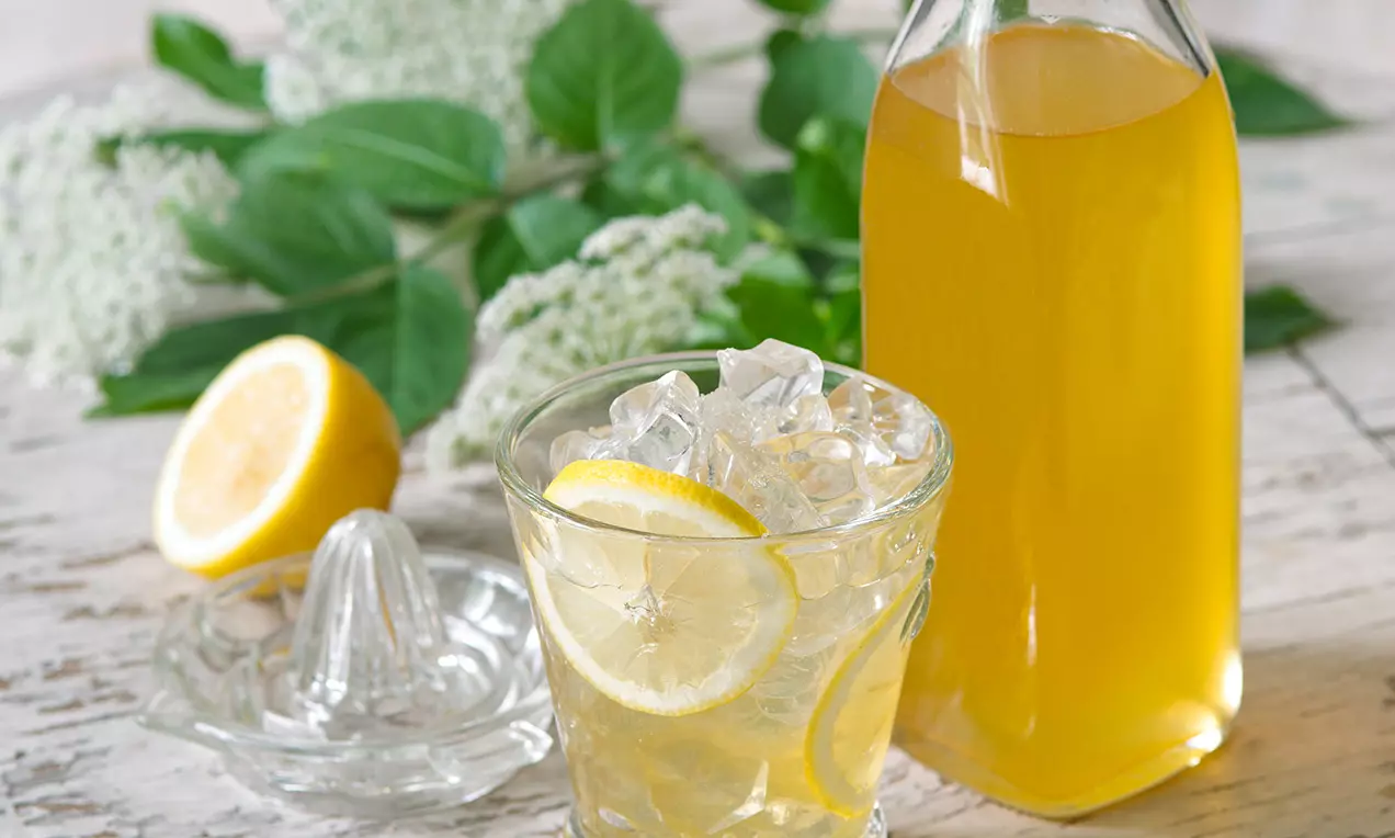 Bezinkový sirup vo vysokej sklenenej fľaši, pripravený s pohárom plným ľadu a plátkami citróna, doplnený listami a kvetmi čiernej bazy