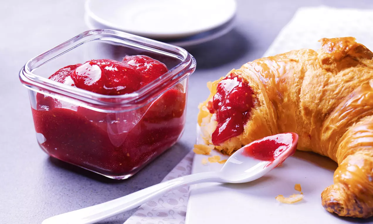 Sklenená nádoba so sýto červeným džemom s jemnou textúrou vedľa zlatistého croissantu a bielej plastovej lyžice na jemne zdobenom obrúsku