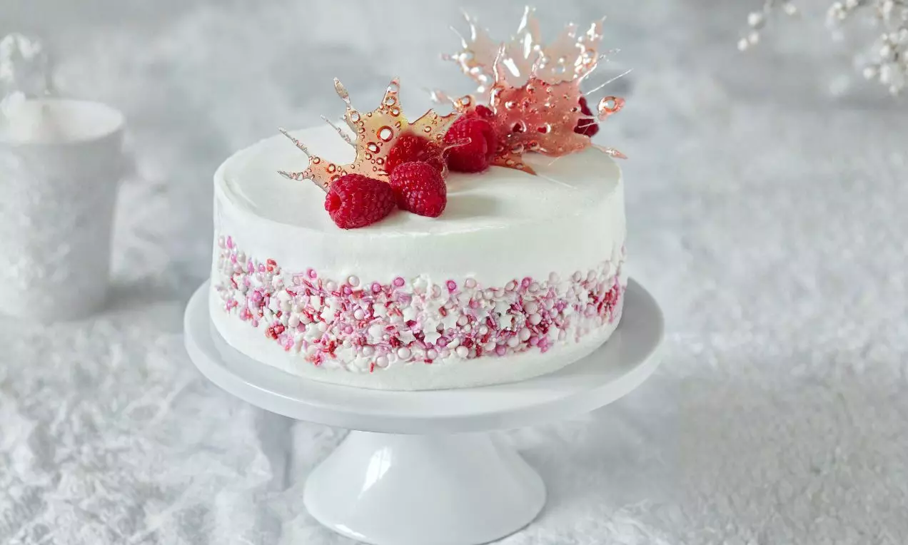 Okrúhly Red Velvet s hladkým bielym krémom, ozdobený malinami, ružovými iskrivými karamelovými dekormi a perlami na porcelánovom podnose