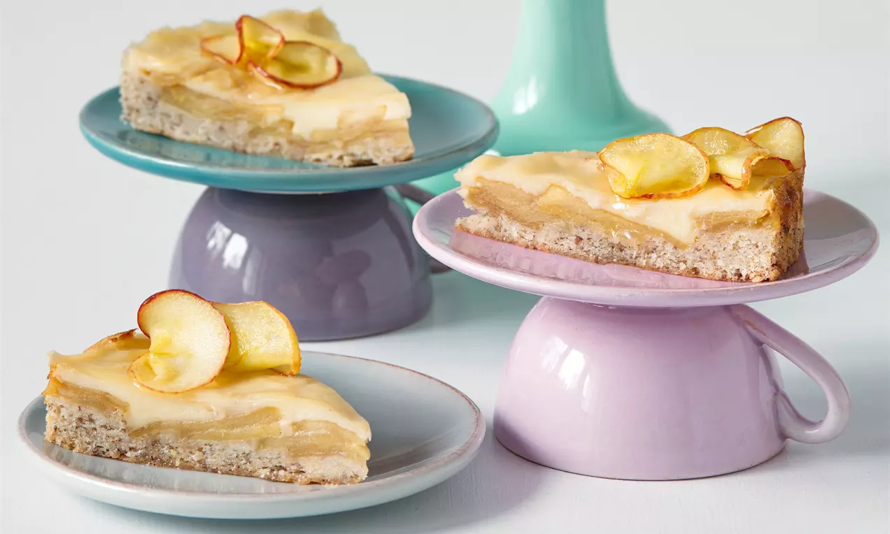 Vegan jablkový koláč s hnedastým základom, krémovou vrstvou a tenkými plátkami jabĺk, podávaný na pastelových tanierikoch