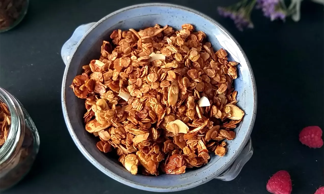 Zlatohnedá medová granola s kúskami kokosu v okrúhlej keramickej miske svetlomodrej farby na tmavom pozadí, doplnená dekoráciou malín a kvetov