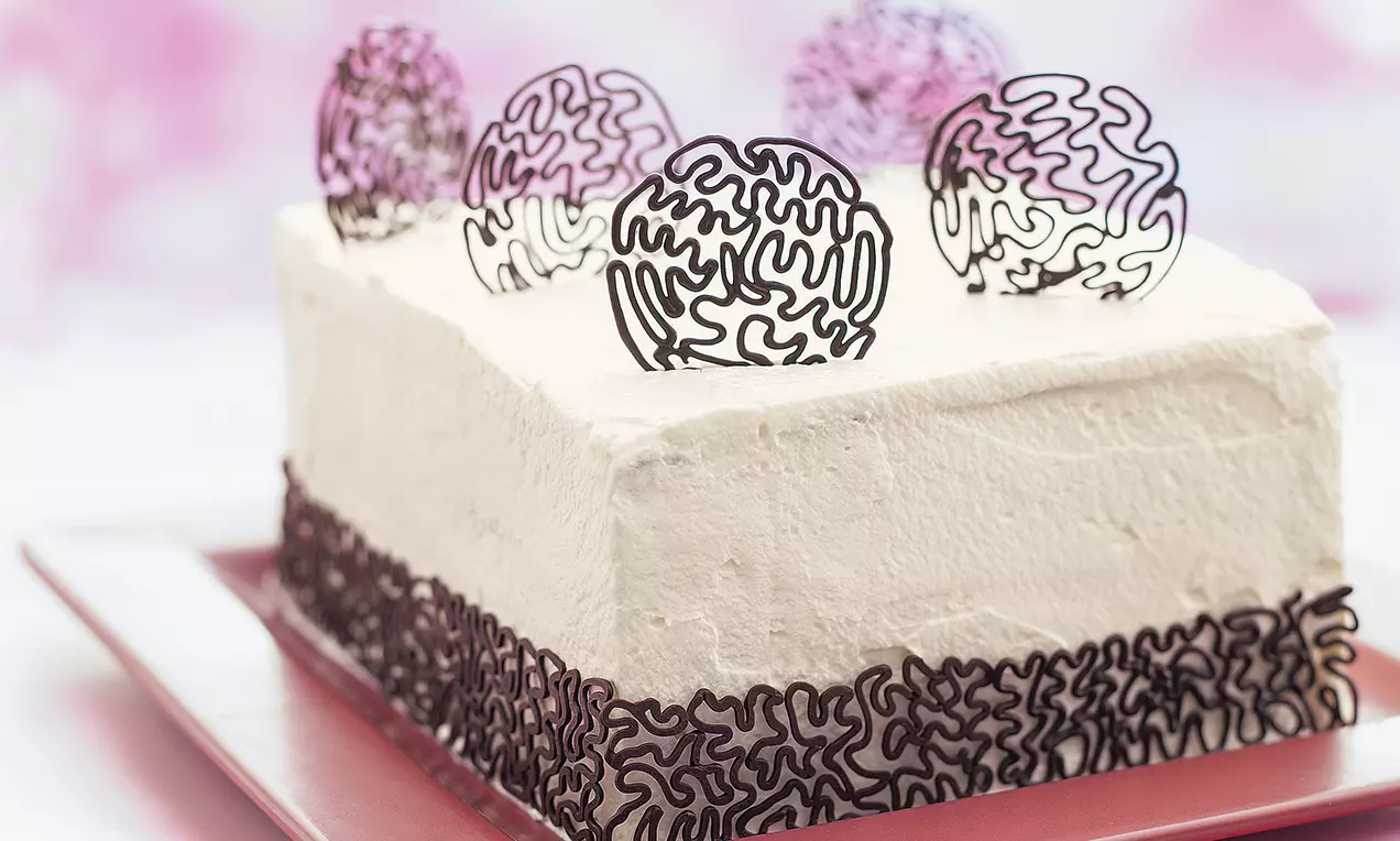 Obdĺžniková torta potiahnutá svetlým krémom zdobená jemnými čokoládovými čipkovanými ornamentmi po obvode a dekoratívnymi čokoládovými ozdobami na vrchnej ploche.