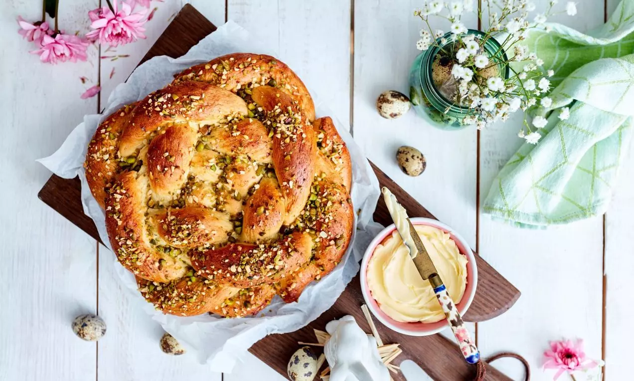 Pletený chlieb zdobený sekanými orechmi na drevenej doske s miskou masla. Okolie zdobené veľkonočnou výzdobou, prepeličími vajíčkami a jarnými kvetmi.