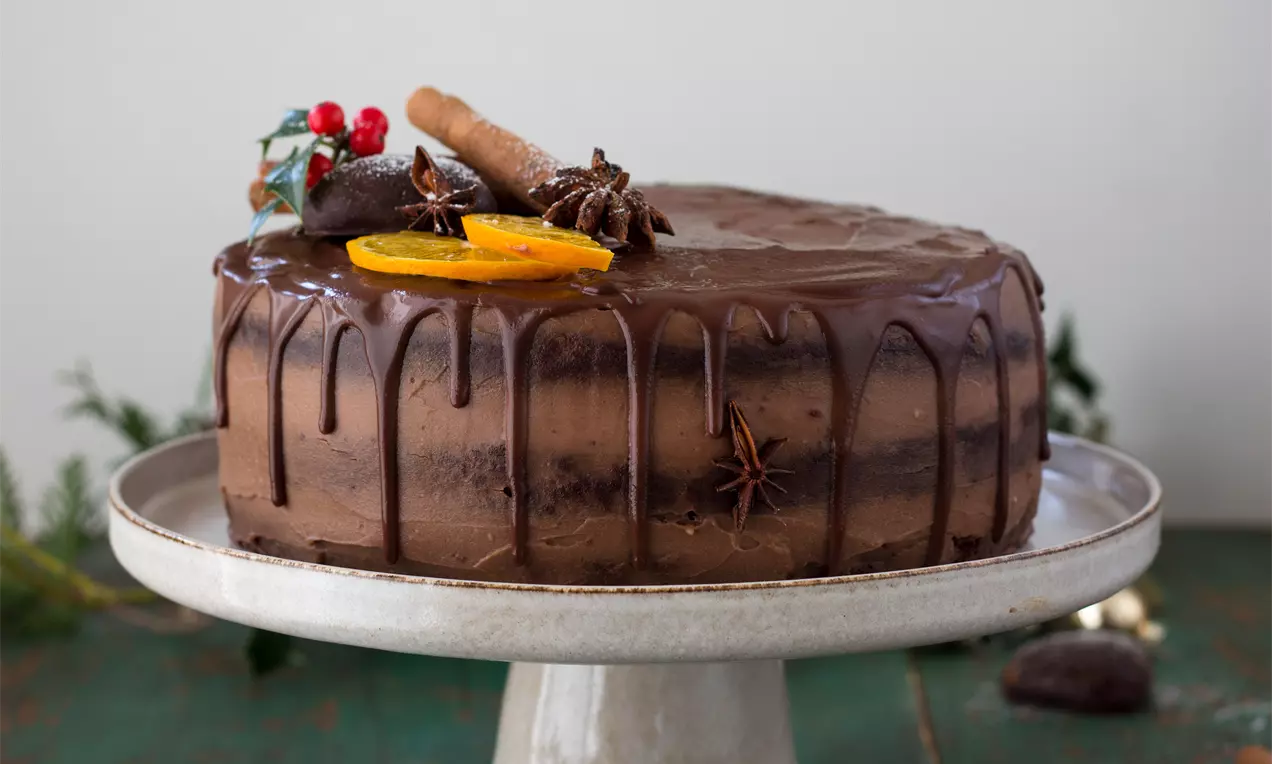 Vznešená čokoládovo-hnedá torta zdobená oranžovými plátkami, hviezdičkami badiánu, červenými bobuľkami a pralinkou na keramickom stojane