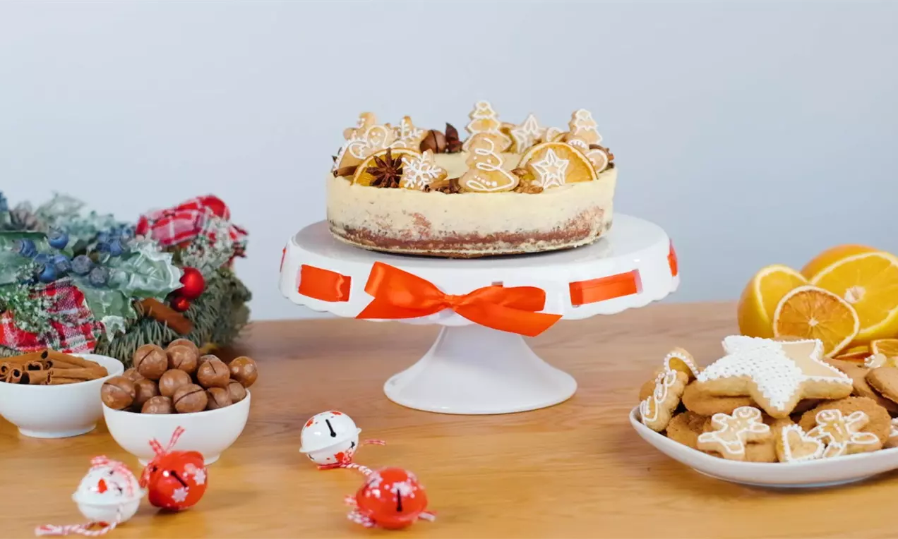 Vysoký krémový cheesecake na bielom stojane s červenou stuhou, ozdobený perníčkami v tvare hviezd a stromčekov s bielou polevou